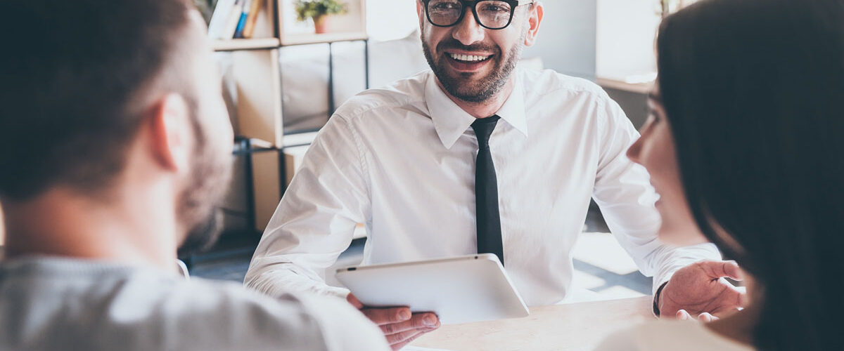 Cheerful mature man in shirt and tie holding digital tablet and gesturing while young couple sitting in front of him at the desk